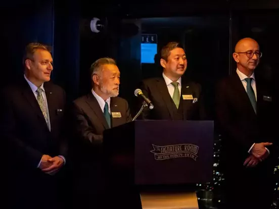 Scott Bradshaw, Kiyotaka Nakao, and Torsten Gilles gathered next to a podium where Makoto Nakao is speaking 